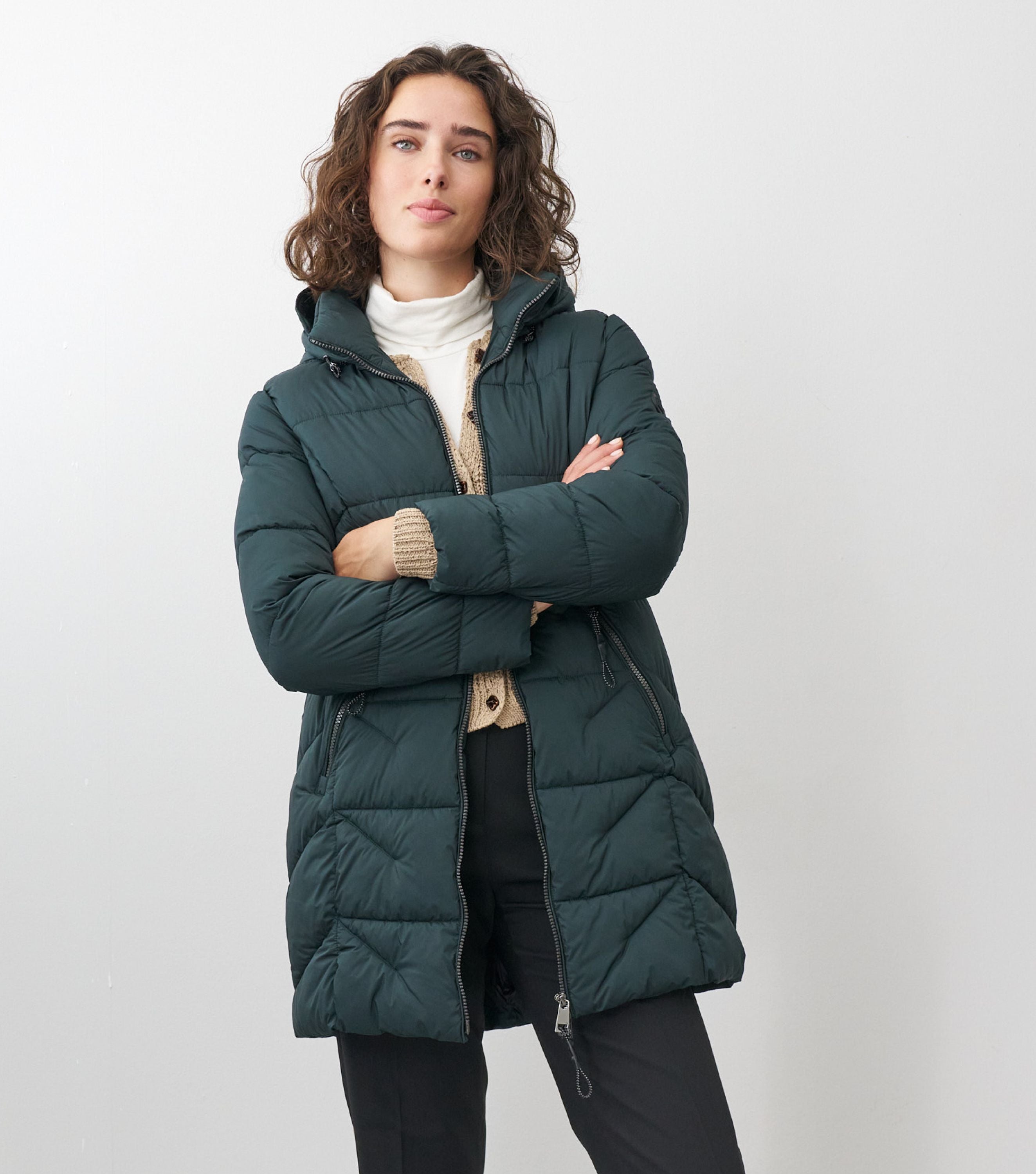 Braunhaarige Frau steht frontal zur Kamera mit verschränkten Armen mit grüner Winterjacke und beigem Sweater drunter