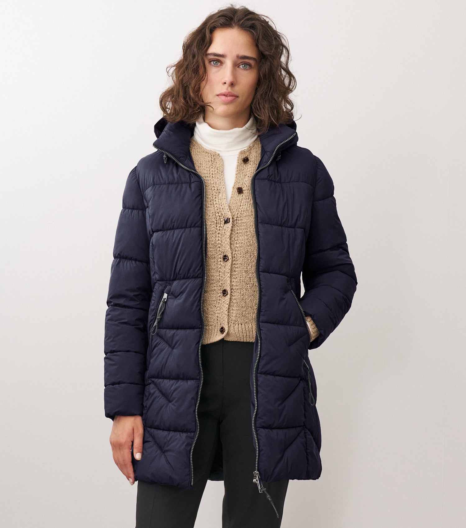 Braunhaarige Frau steht frontal zur Kamera mit blauer Winterjacke und beigem Sweater drunter 