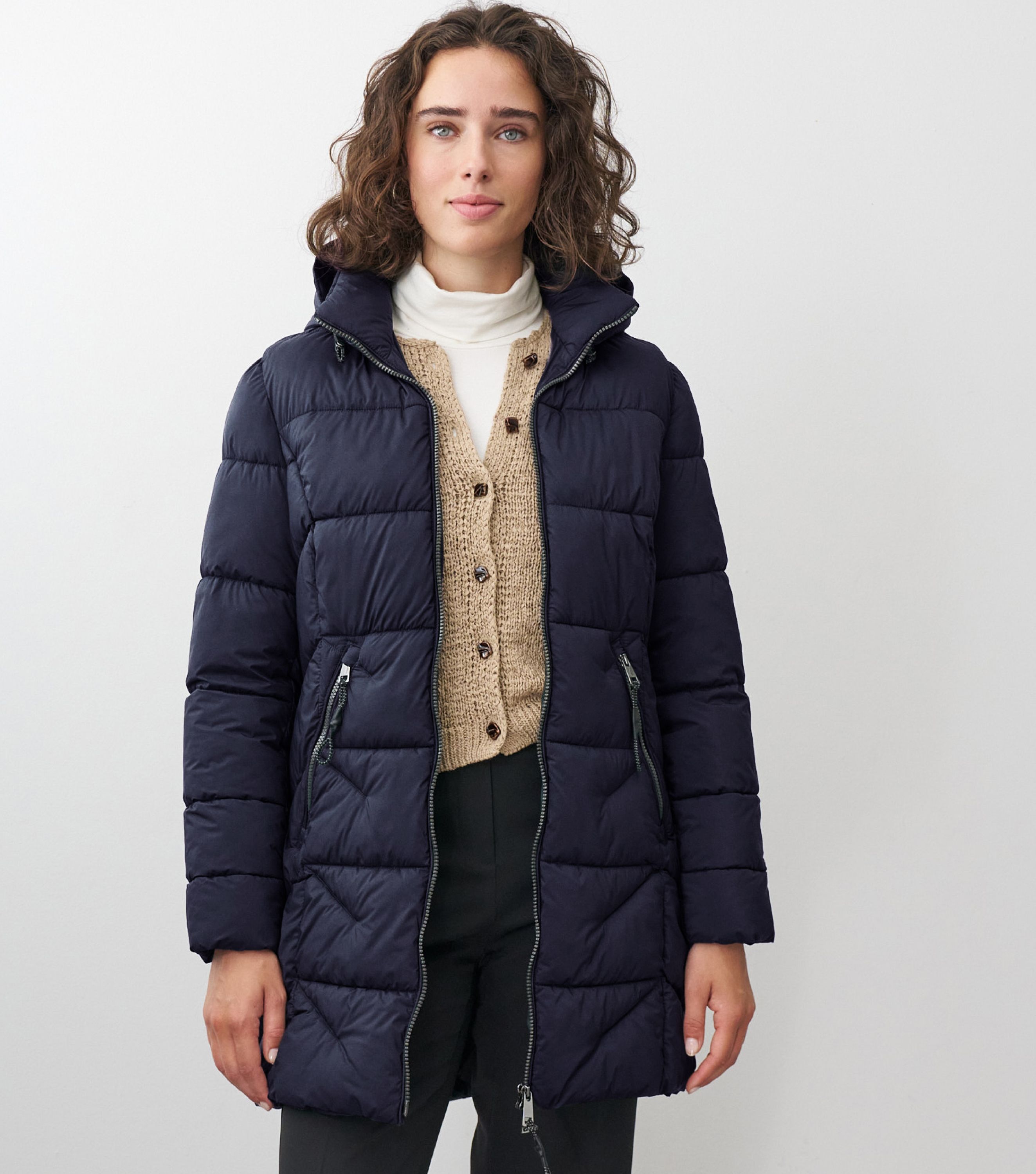 Braunhaarige Frau steht frontal zur Kamera mit blauer Winterjacke und beigem Sweater drunter 